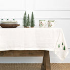 Table setting with a white tablecloth featuring green tree patterns, mugs, and small Christmas trees on a white wall background.