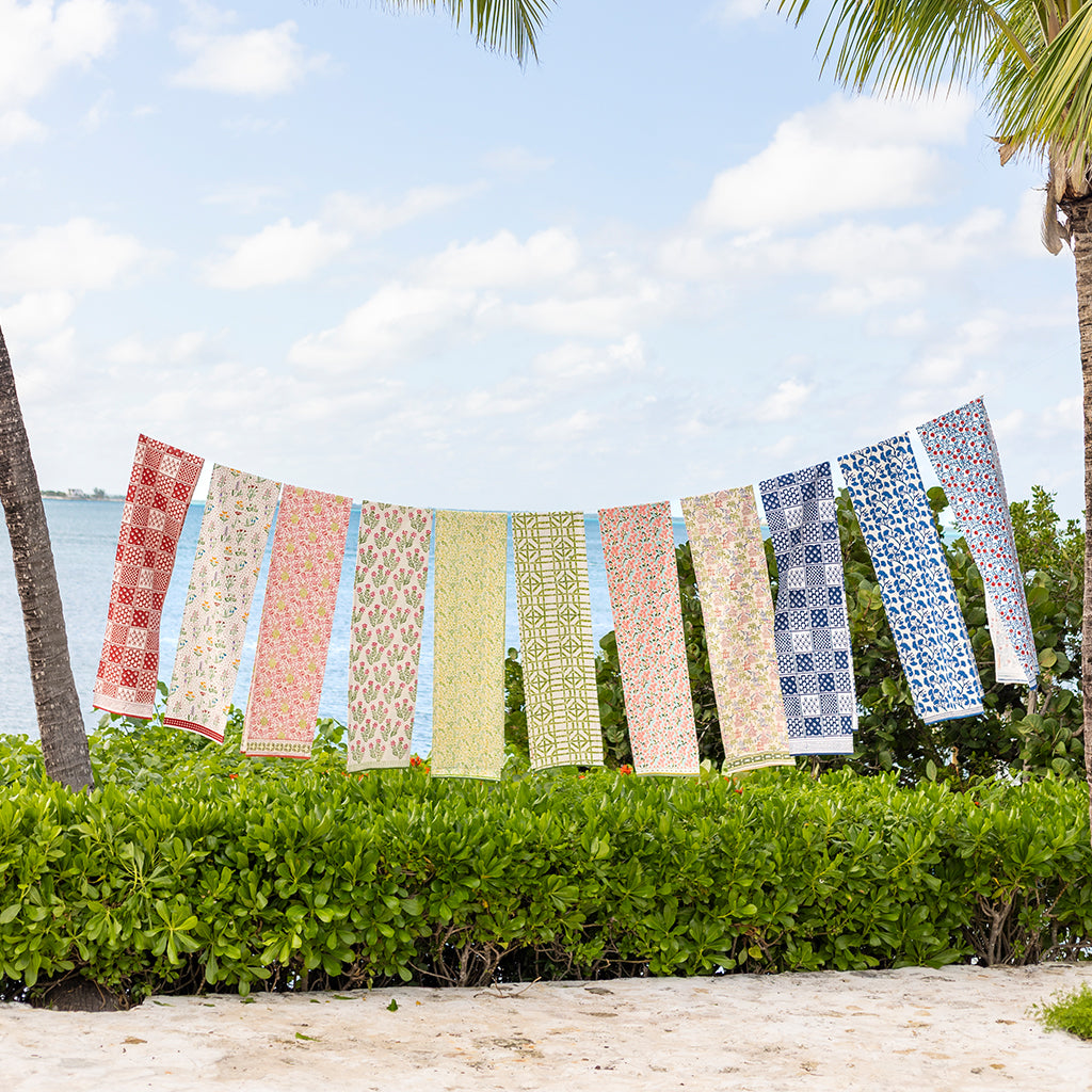 Colorful patterned scarves hanging on a line against a blue sky with clouds and greenery.