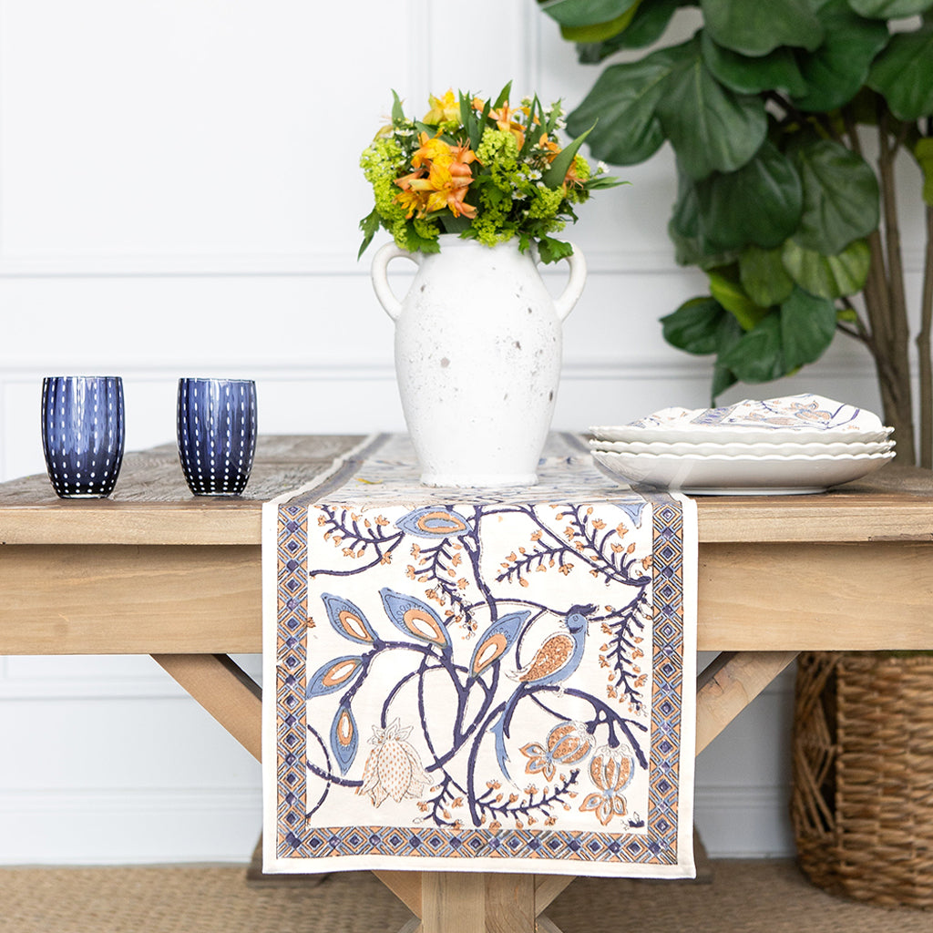 Decorative table runner with floral and bird pattern on a wooden table with flowers and glasses.