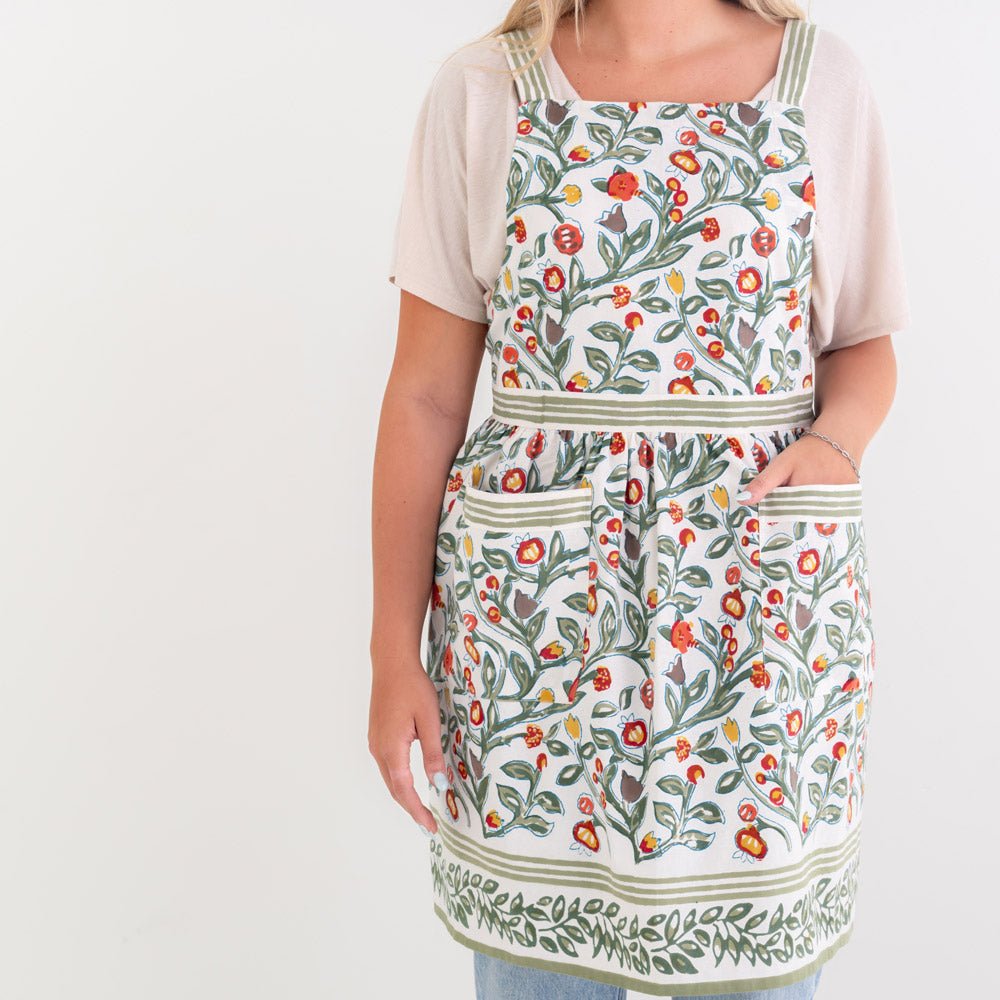 model wearing apron printed with green vines and floral buds in shades of crimson, marigold and deep orange