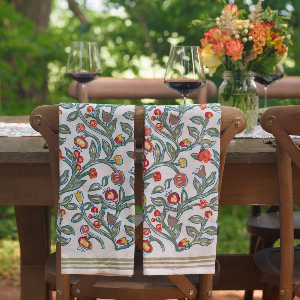 two tea towels with floral pattern in shades of sage green, crimson, marigold and deep orange hanging over the back of a wooden chair