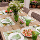 Outdoor table with placemats, napkins, green glasses, florals, and food. 