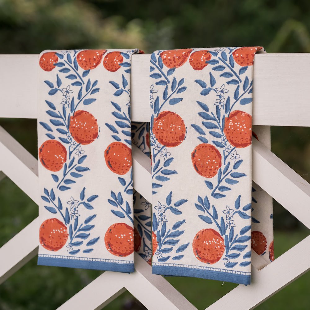 two tea towels printed with oranges and blue floral design hanging from white fence